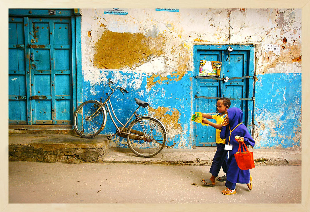 A photograph capturing two children walking on a street in Stone Town, Zanzibar with vibrant colored walls and a bicycle parked against one of the walls. The individuals appear to be engaging with each other, and the setting has a casual, exploratory vibe.