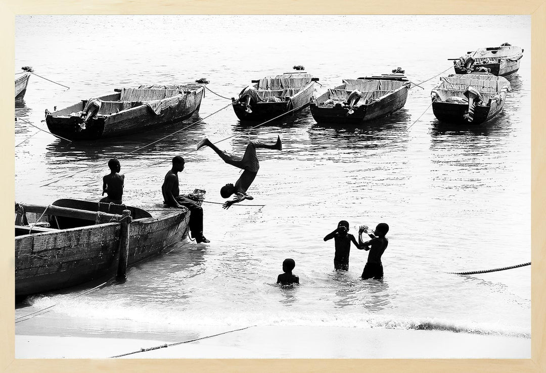 Black and white photograph of young boys on a beach with boats in the background, capturing a carefree moment.