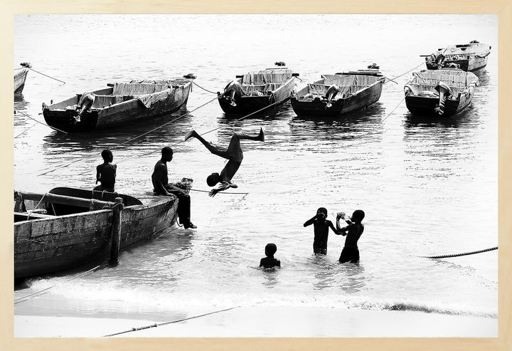 Black and white photograph of young boys on a beach with boats in the background, capturing a carefree moment.